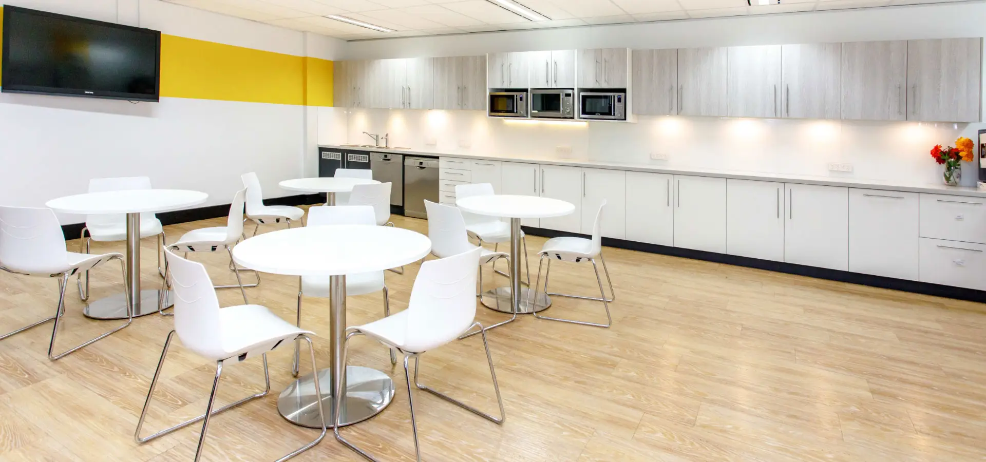 Modern office break room with white tables and chairs, kitchenette with white cabinets, and a yellow accent wall.