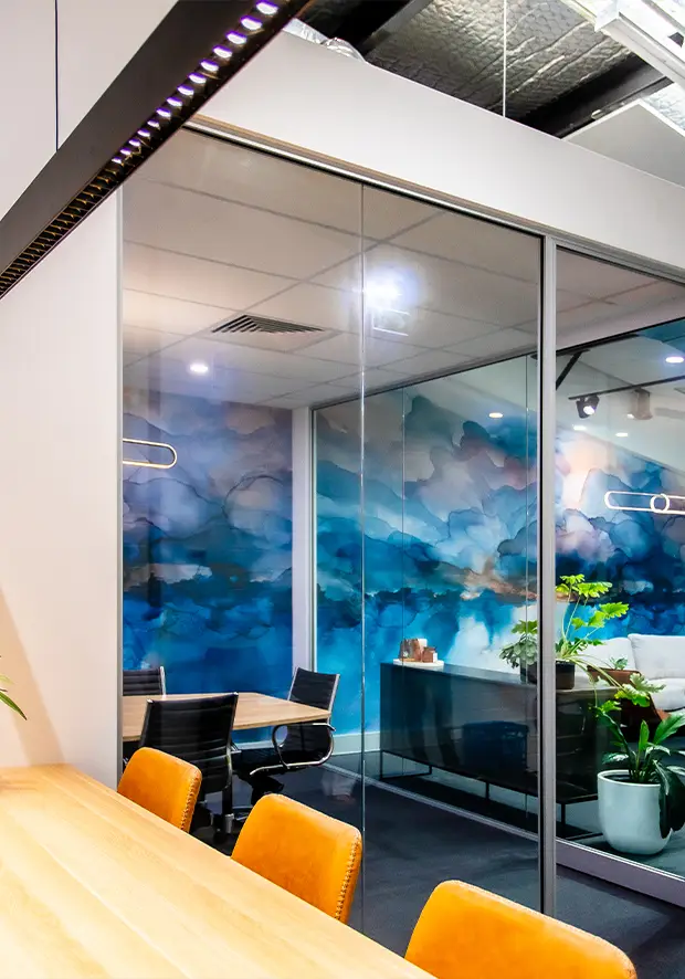 Modern office meeting room with glass walls, featuring a large abstract blue and white wall mural, black chairs with orange cushions, a wooden table in the foreground, and a potted plant.