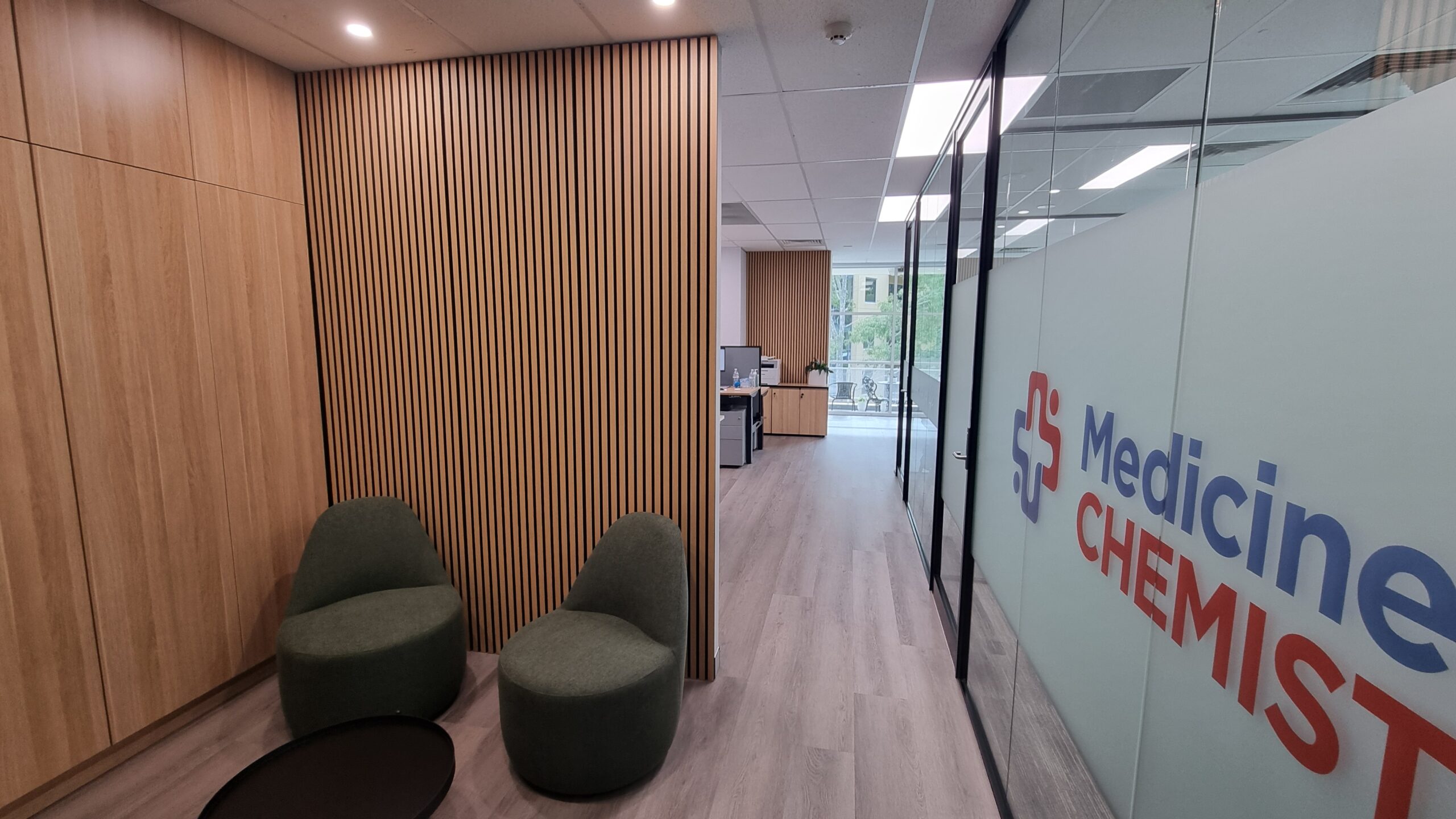 Reception and waiting area at Medicines R Us in Norwest Business Park featuring timber slat wall panelling, glass office partitions with branded frosting, modern seating, and timber-look flooring.